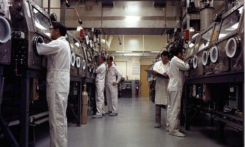 photo of several scientists in lab coveralls standing in front of two rows of glove boxes, with their hands inside the gloves so they can work with plutonium